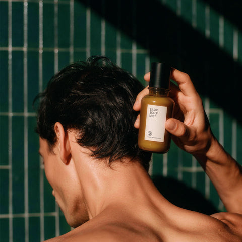 A man with dark hair is shown from behind, holding a bottle of Baseline Skincare 'Basic Scalp Mist' against a dark green tiled wall.
