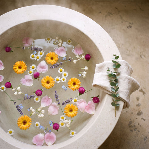 An overhead view of a warm bath strewn with seven auspicious flowers — calendula, chamomile, lavender, jasmine, forget-me-not, globe amaranth, and peony — for a Seven Flowers ritual bath