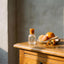 A small frosted glass bottle with a cork stopper sits on a wooden cabinet beside a plate of oranges and orange peels, against a textured gray wall.